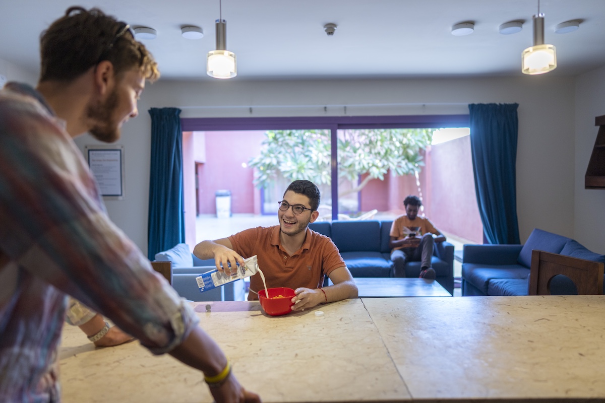 Students socialize in a residence hall common area while one prepares cereal in the shared kitchen.
