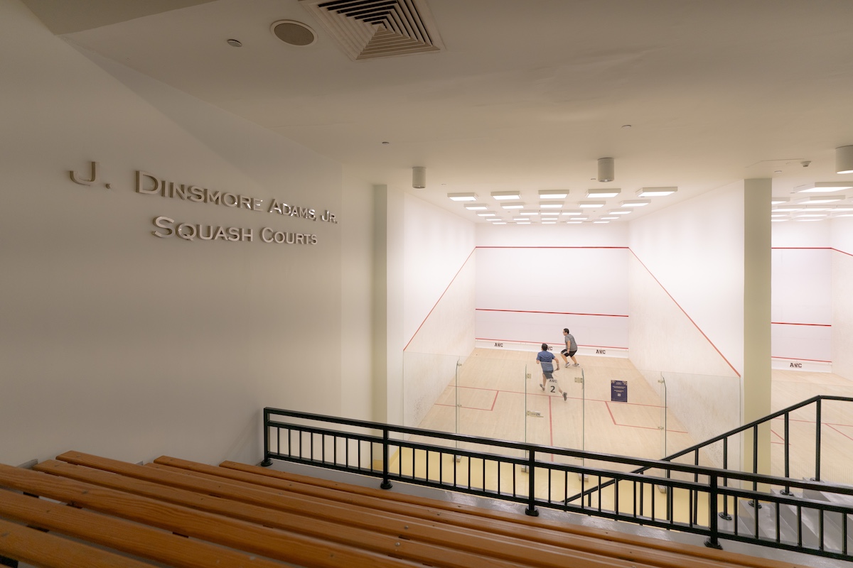 Two players compete in a squash match inside the J. Dinsmore Adams Jr. Squash Courts facility at AUC, viewed from spectator seating.