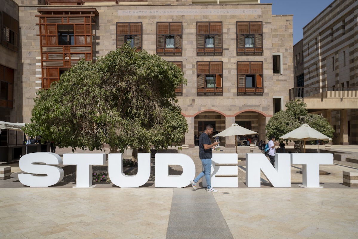Large &ldquo;STUDENT&rdquo; sign in the courtyard of the Hatem and Janet Mostafa Core Academic Center with students walking nearby.