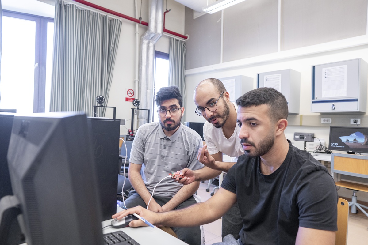 Three males are looking into a computer screen. They are in a classroom