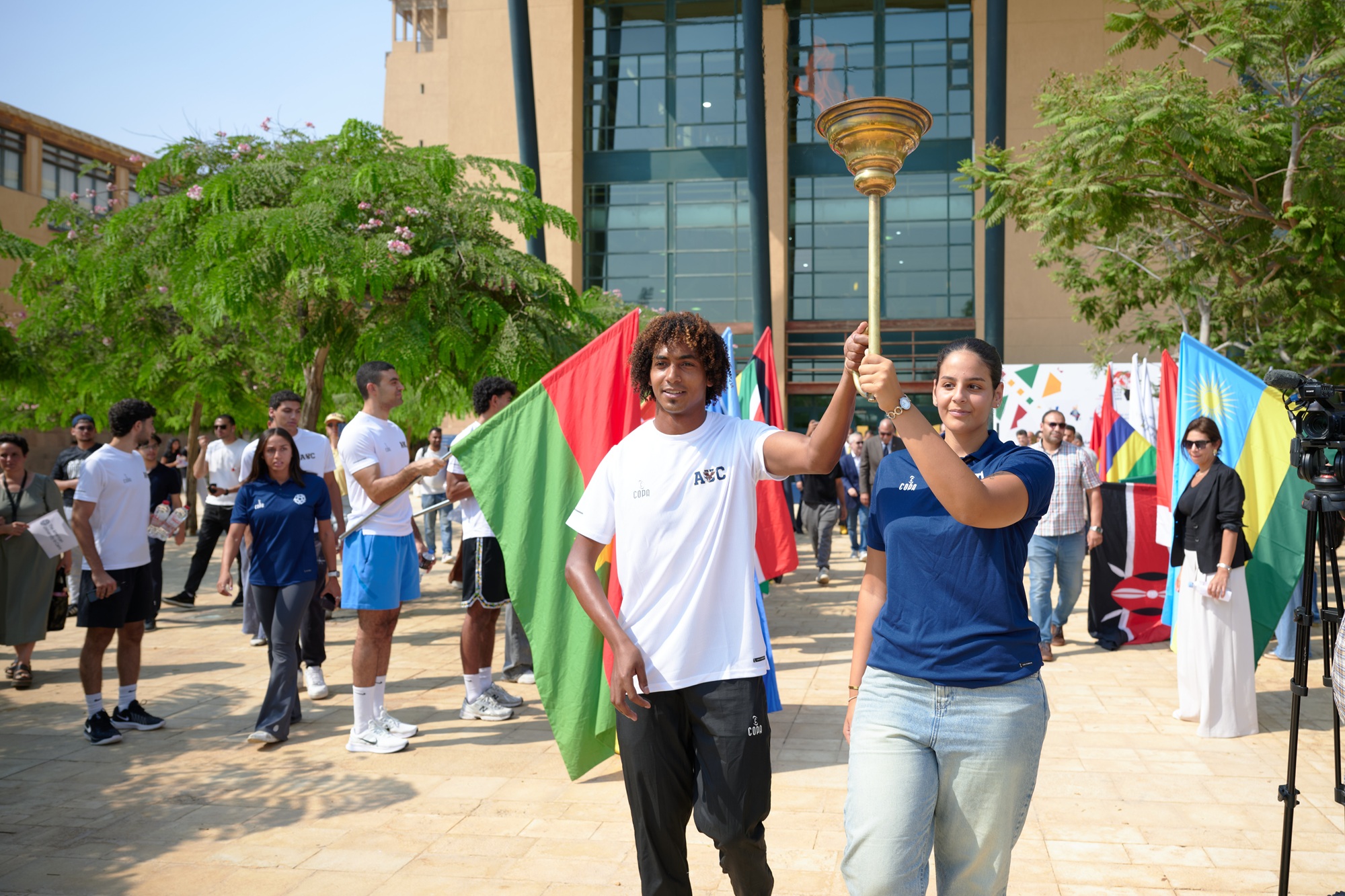 A male and a female are holding a torch. There are people around them holding country flags