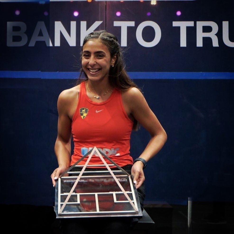 Hania El Hammamy stands smiling on a court while holding a squash trophy