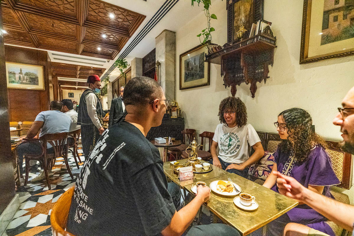 Abdelhalim smiles while sitting with his friends in a cafe in Khan al Kalili. There is a gold colored coffee set in front of him.