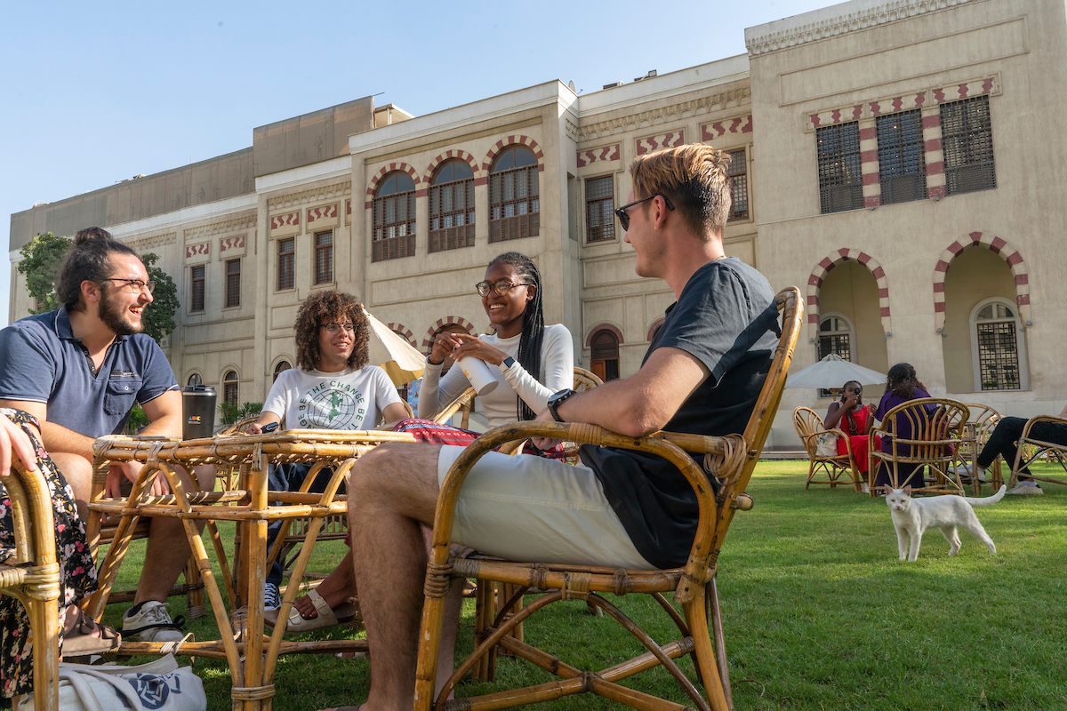 Abdelhalim sits with three friends at a wicker table at AUC Tahrir campus. There is a small white cat in the bottom corner. 