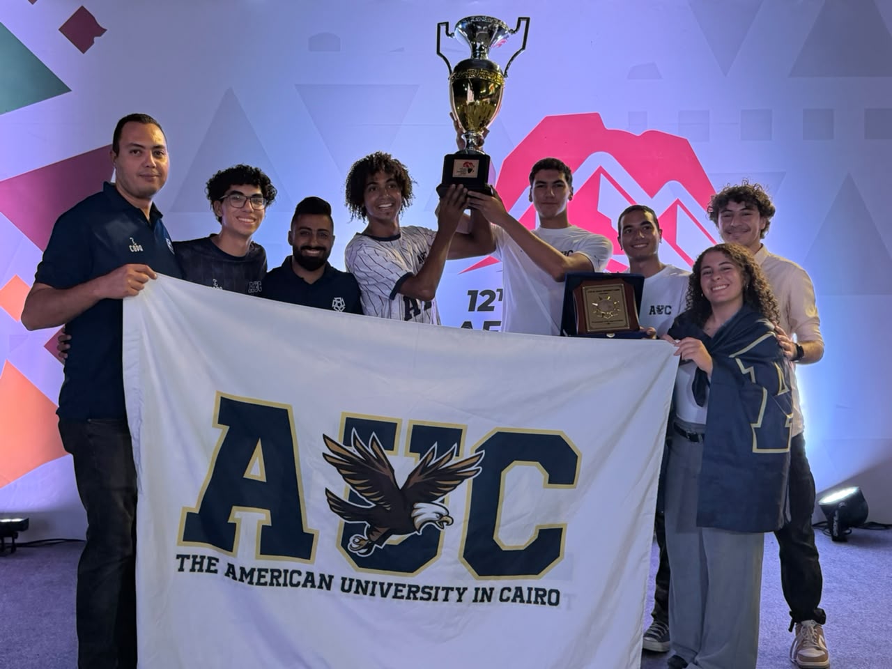 Students and coaches holding up an AUC banner and trophy at the closing ceremony.