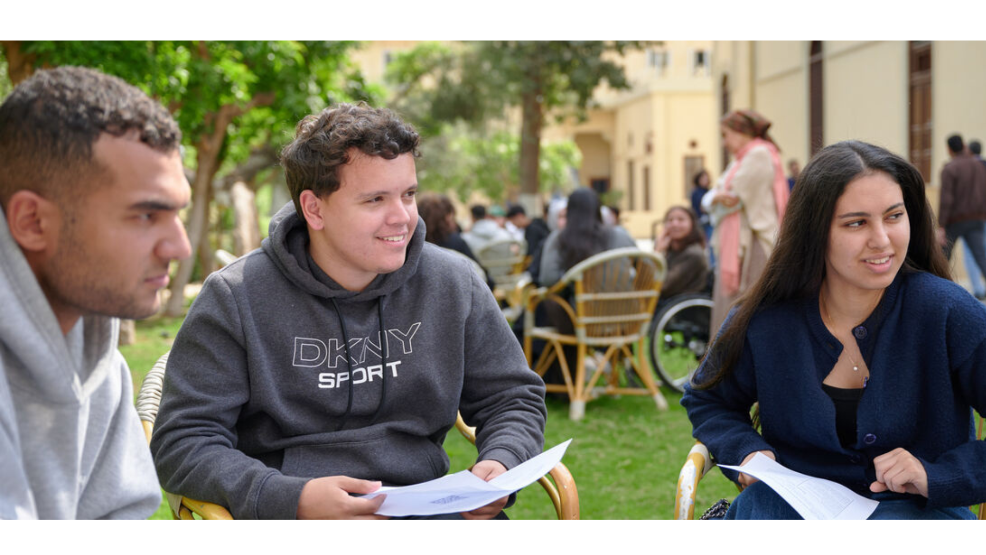 Three students seated outside listen attentively and and smile at a speaker