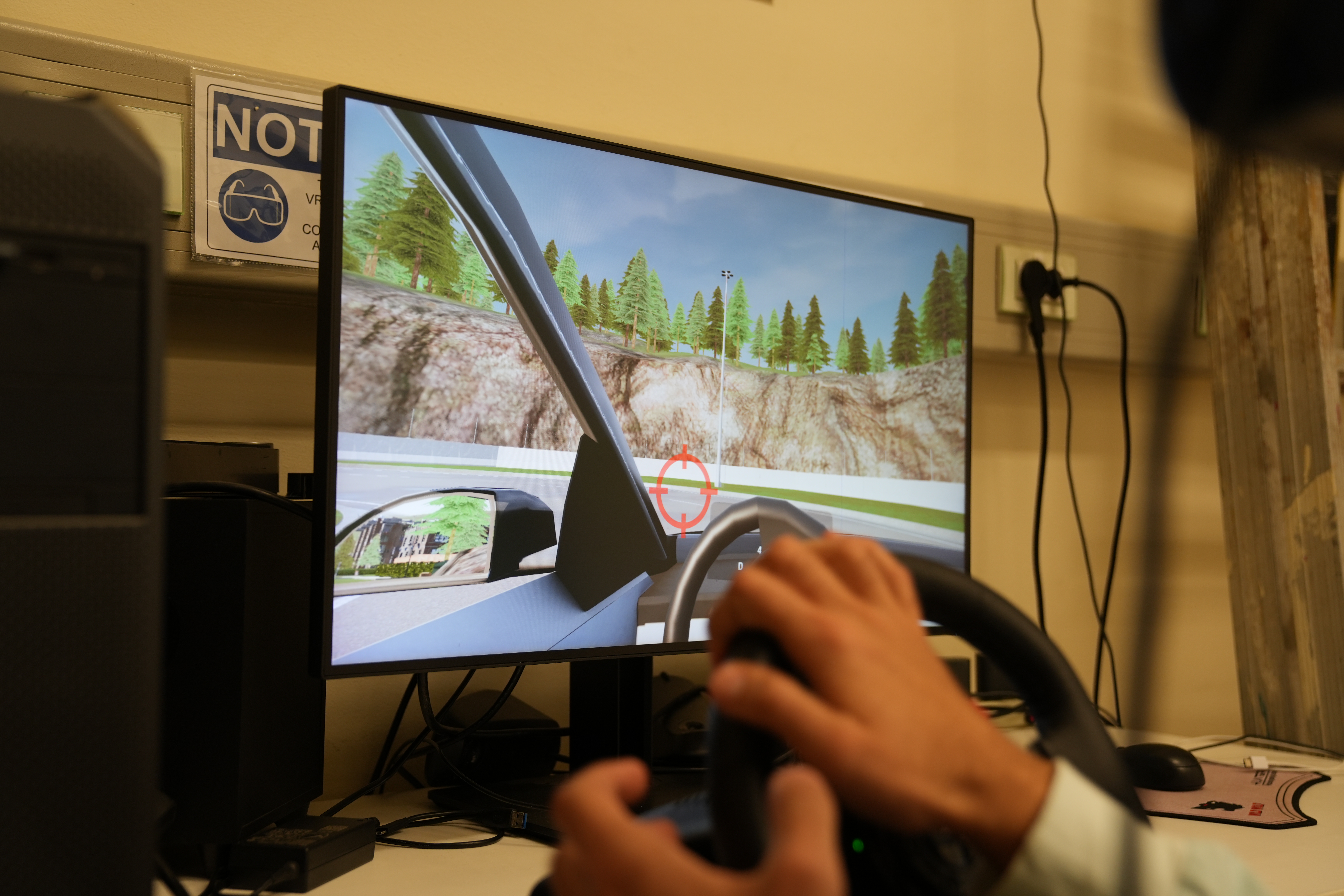 A person wearing a virtual reality headset turns a wheel and a monitor displaying a virtual car and road turns as well