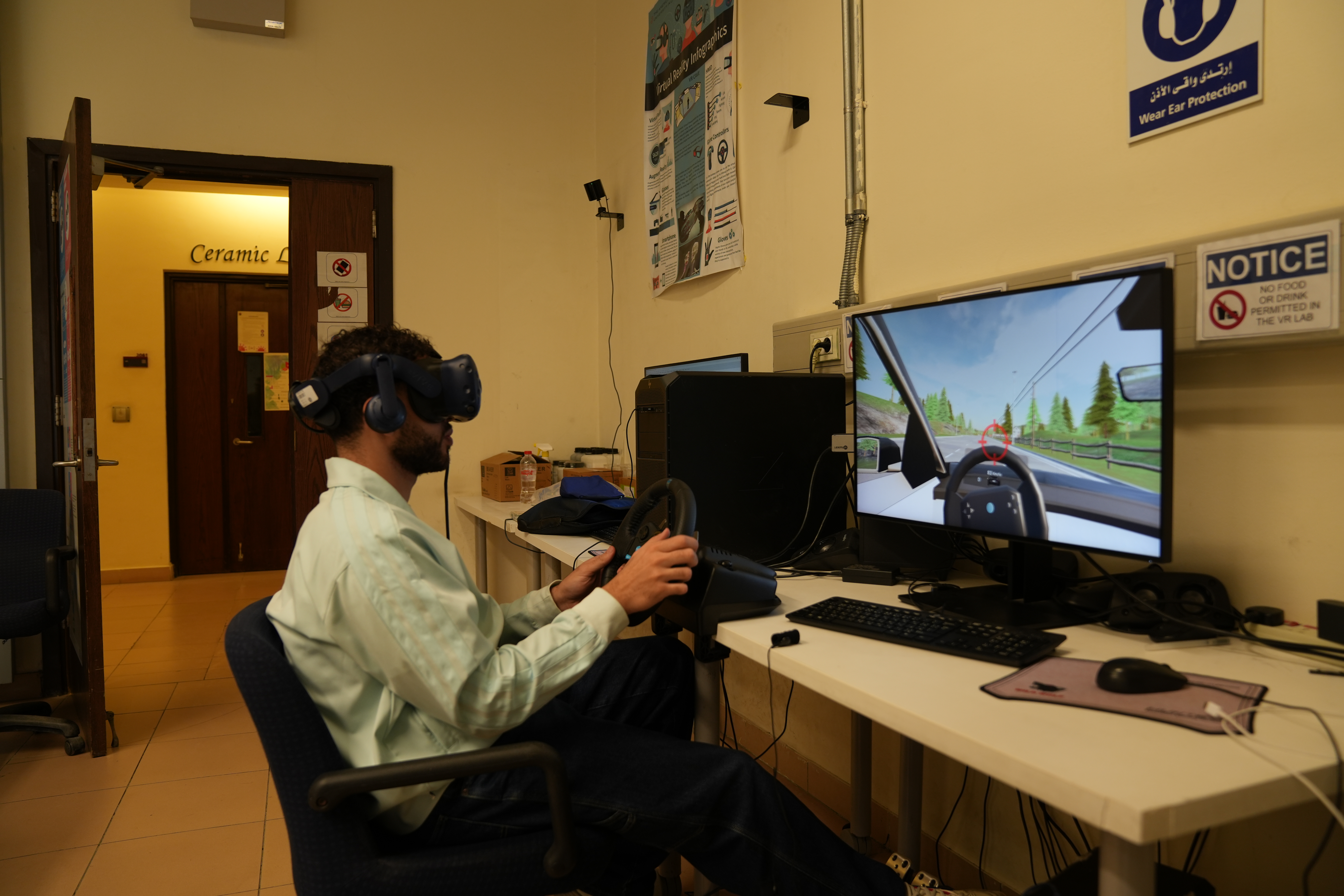 A research assistant wearing a virtual reality headset turns a steering wheel in front of a monitor which displays a virtual car turning