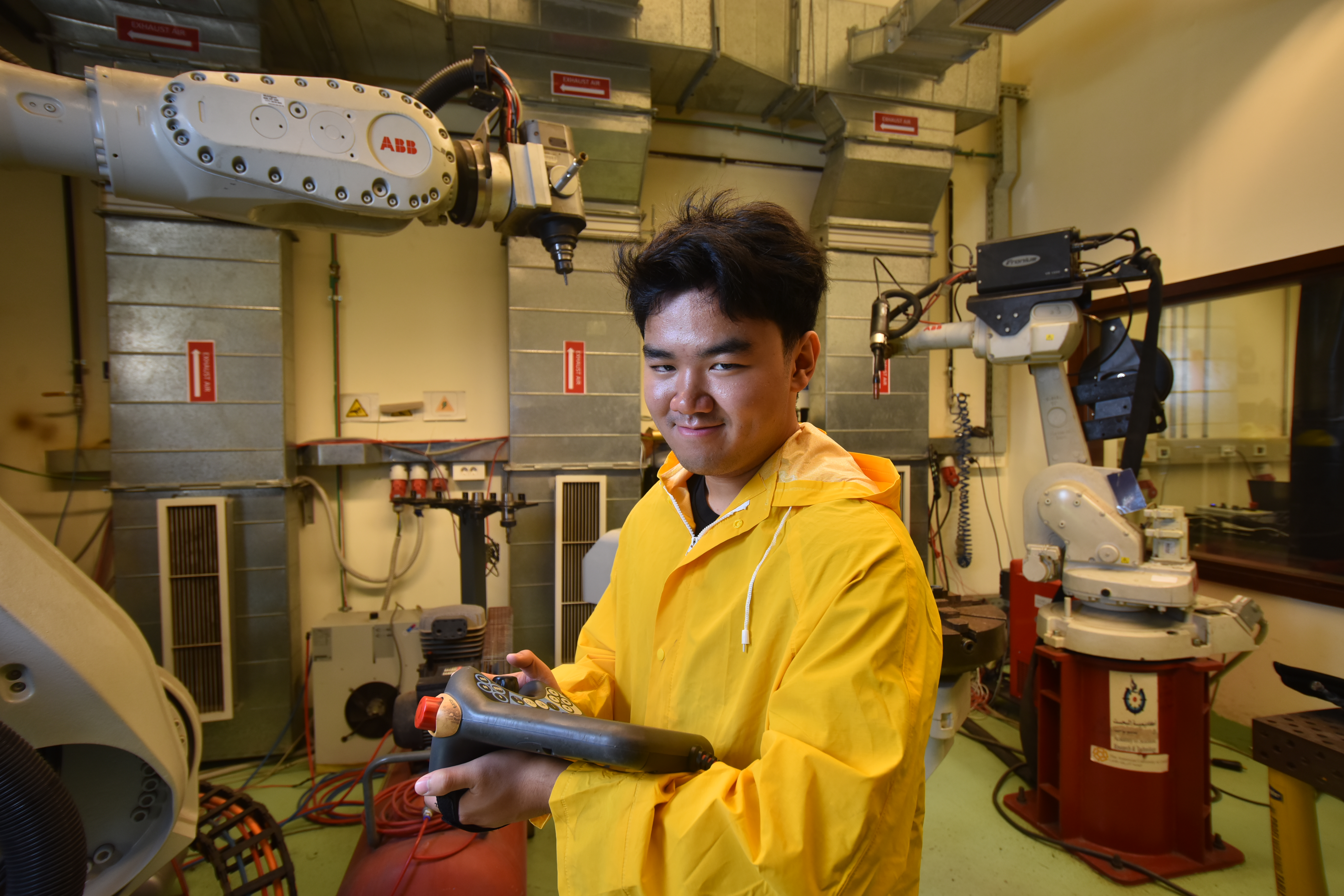Anthony Nguyen holds a tablet in a mechanical workshop with robotic arms behind him