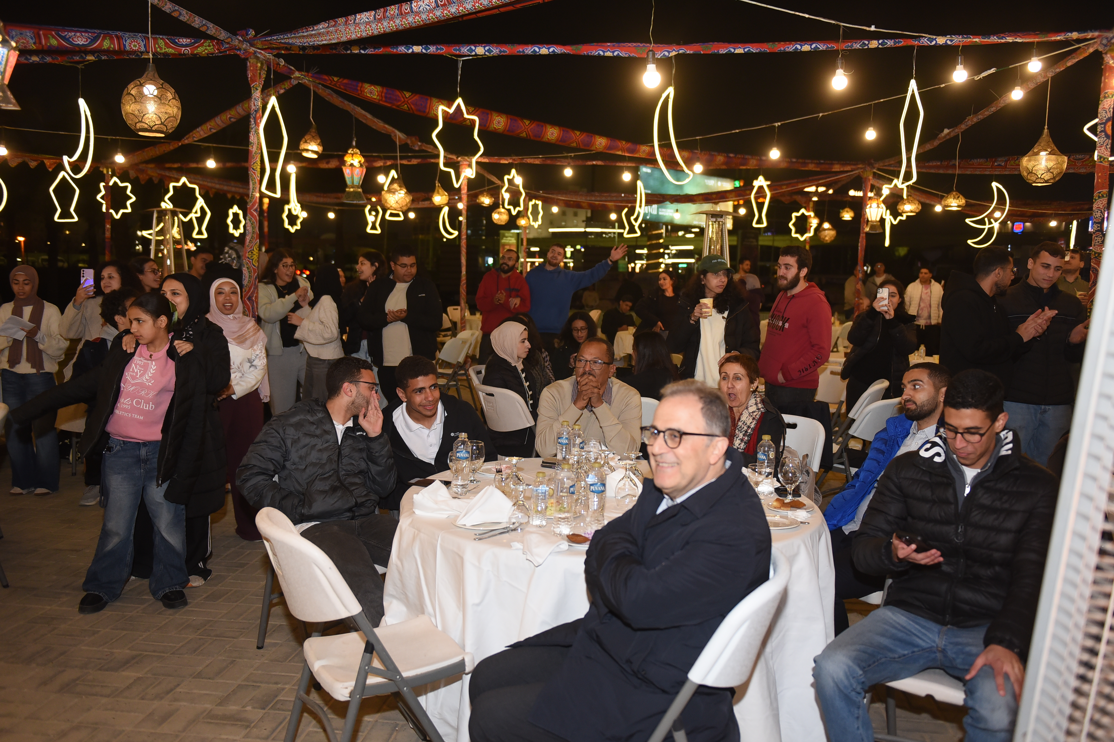 A group of people sit outside underneath crescent and lantern shaped lights