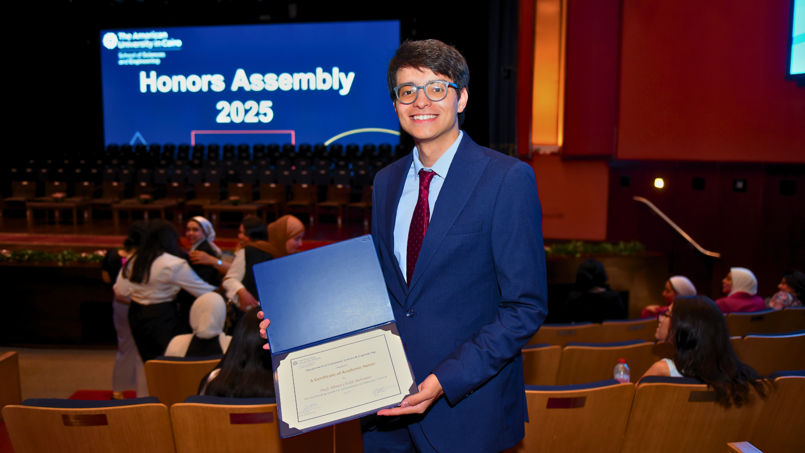 Dahmane smiles in a theater, holding up a certificate with a screen on stage that reads "Honors Assembly 2025"