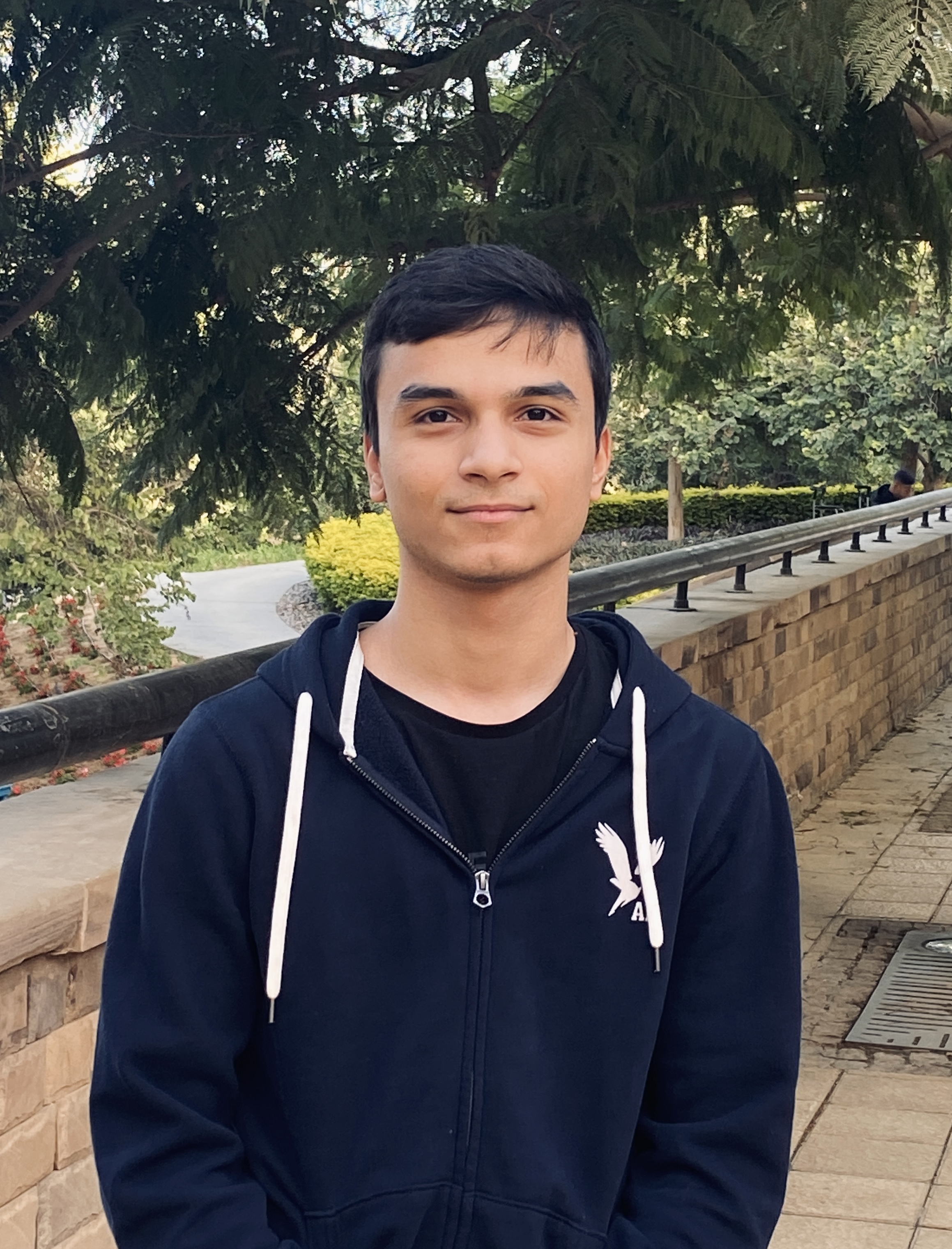 A young man with short dark hair stands in front of trees on a college campus wearing a black hoodie.