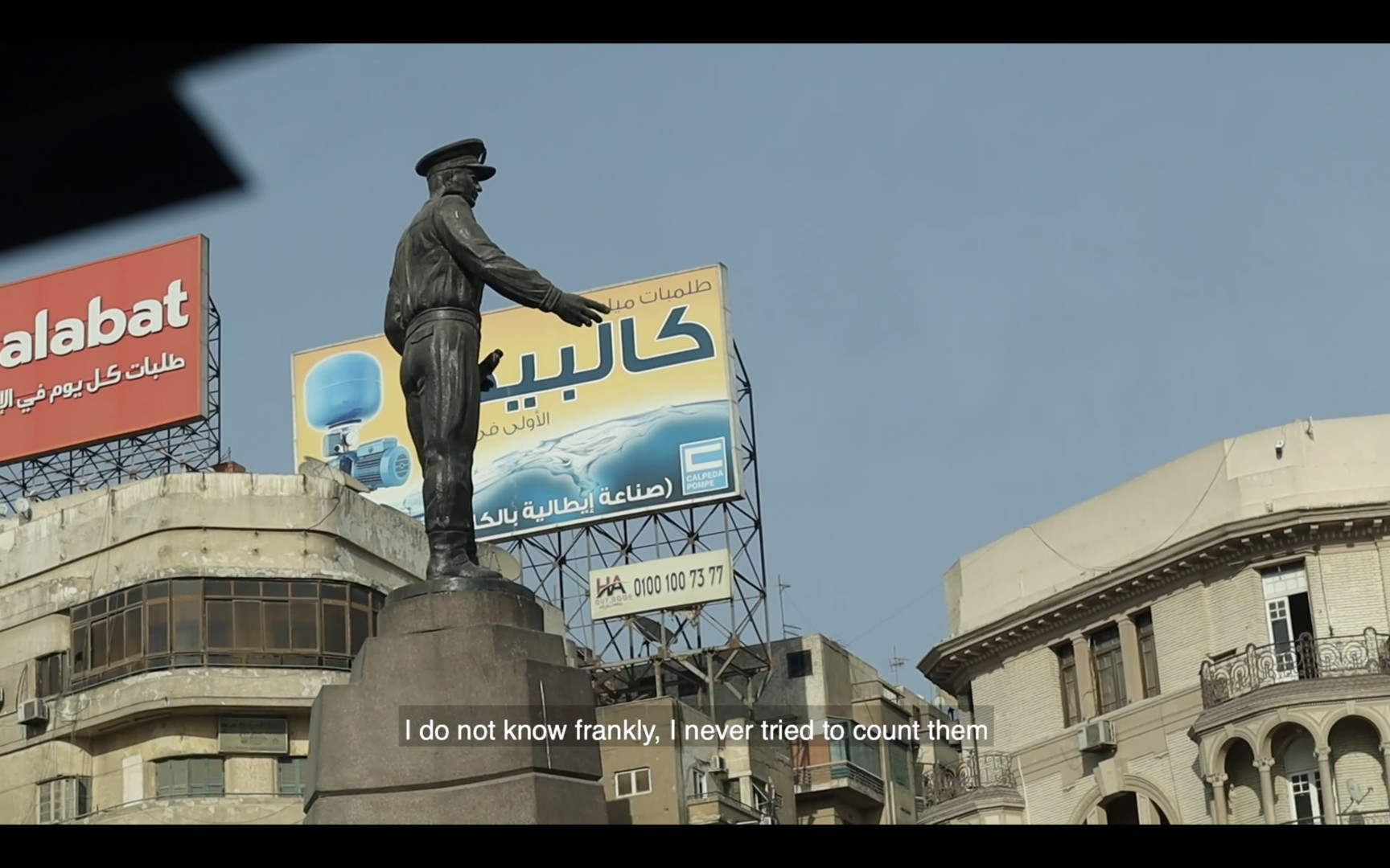 A screenshot of the film "Way Home". The shot shows a statue in the center of a square with buildings and billboards behind it. Caption at the bottom read "I do not know frankly, I never tried to count them."