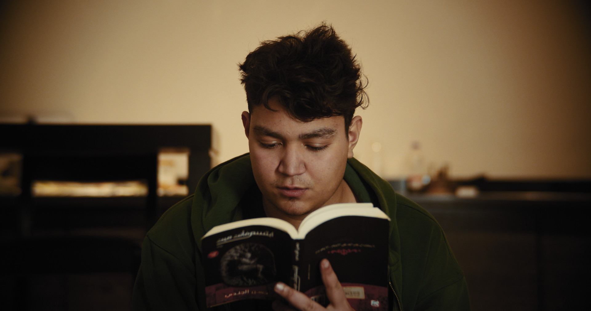 A screenshot of the film "Okay." A young man sits in the center of the screen while reading a book. The lighting and tone are dark and ominous.