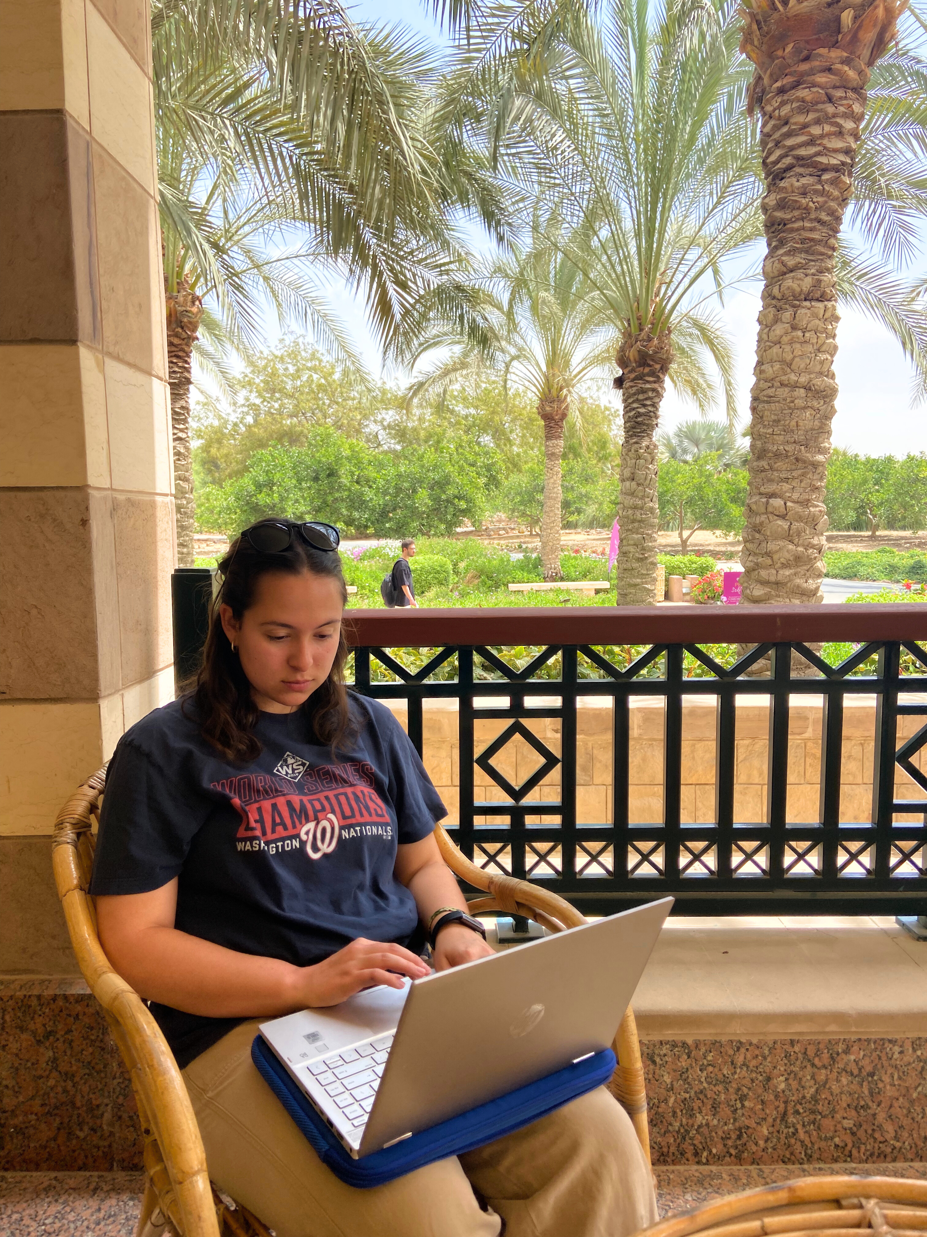 McDermott sits at a table behind the administration building, working on a laptop. The garden is visible behind her and there is a young man walking in the background.  