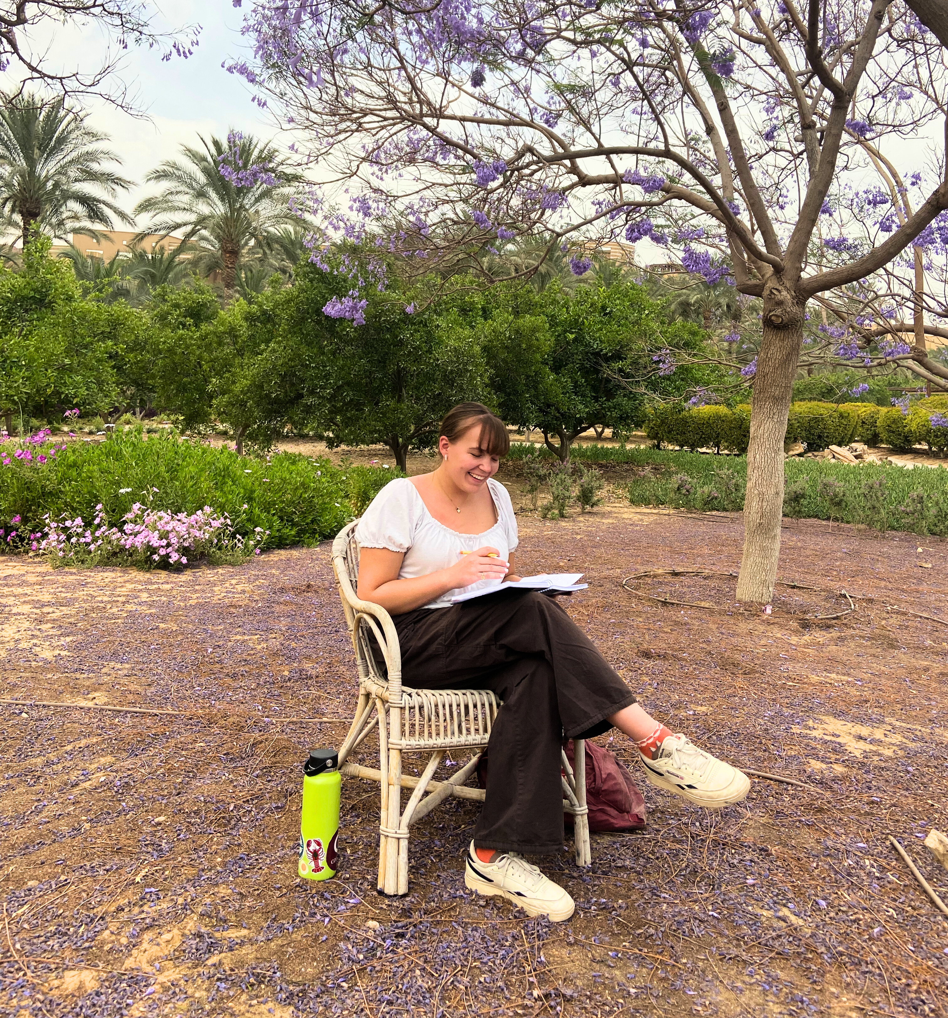 Corson sits under two flowering purple trees in the garden. She is writing in a notebook and there is a green water bottle near her chair. The rest of the garden and the Administration Building is visible in the background. 