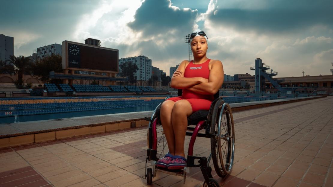 Aya Abbas '23 sits in a wheel chair wearing a swimsuit, swim cap and goggles in front of the AUC pool with her arms crossed, looking determined 