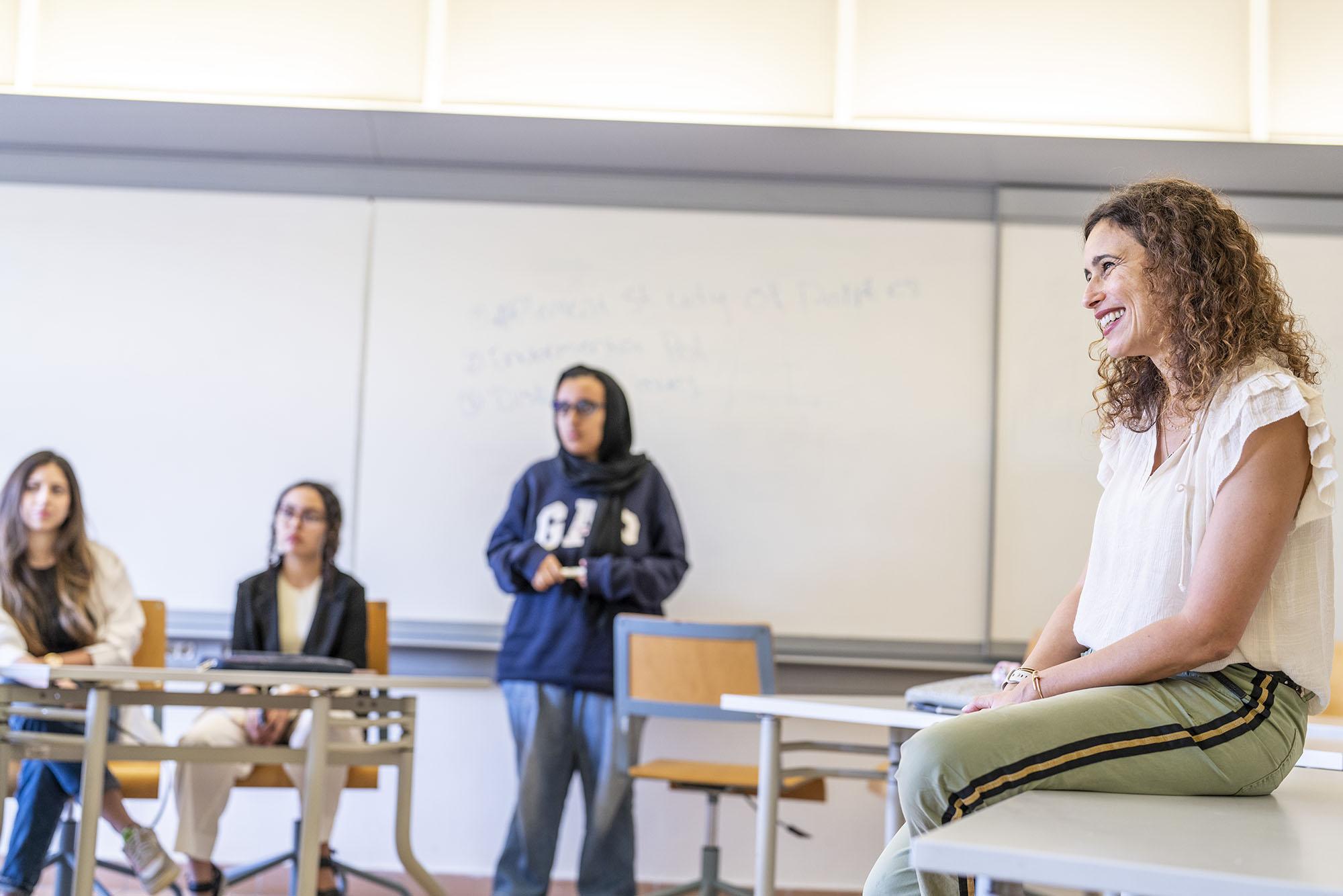 Students in class and faculty sitting on desk smiling looking at students