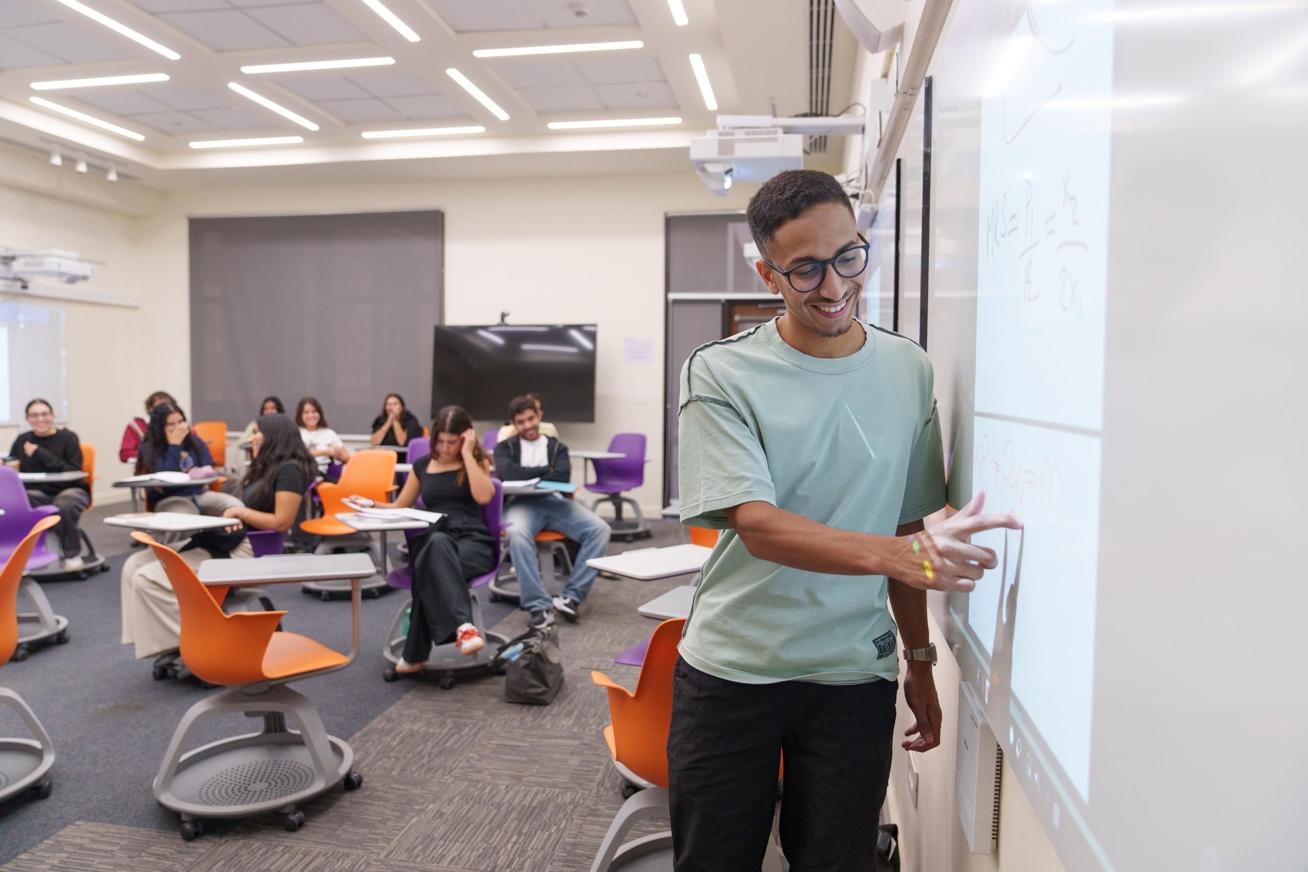a boy wearing a mint tsirt and eye glasses writing on the white board in class