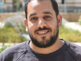 A headshot of a man smiling wearing a black tshirt in an outdoor setting