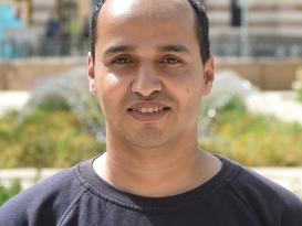 A headshot of a man smiling wearing a black tshirt in an outdoor setting