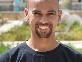 A headshot of a man smiling wearing a black tshirt in an outdoor setting