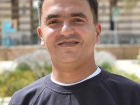 A headshot of a man smiling wearing a black tshirt in an outdoor setting