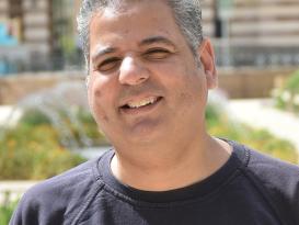 A headshot of a man smiling wearing a black tshirt in an outdoor setting