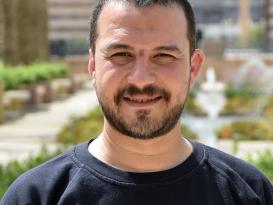 A headshot of a man smiling wearing a black tshirt in an outdoor setting