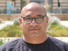 A headshot of a man smiling wearing a black tshirt in an outdoor setting