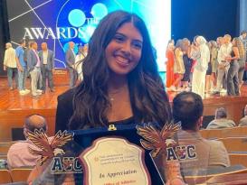 A female is smiling and holding three trophies. Text: In appreciation. Office of Athletics. The letters A U C are written on them and there is an eagle next to the letters. There are people around her