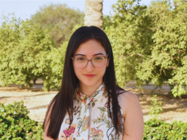 A female is smiling and standing in a garden. She is wearing glasses