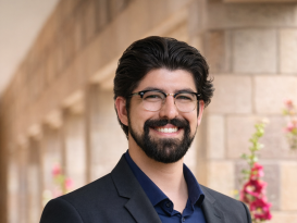 A man is standing and smiling. He is wearing glasses and a suit