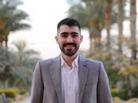 a headshot of a man with short black hair and black beard wearing a grey suit