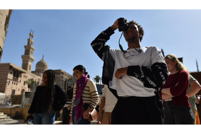 A student holds a camera to his eye while photographing something; a group of students stands behind him in conversation