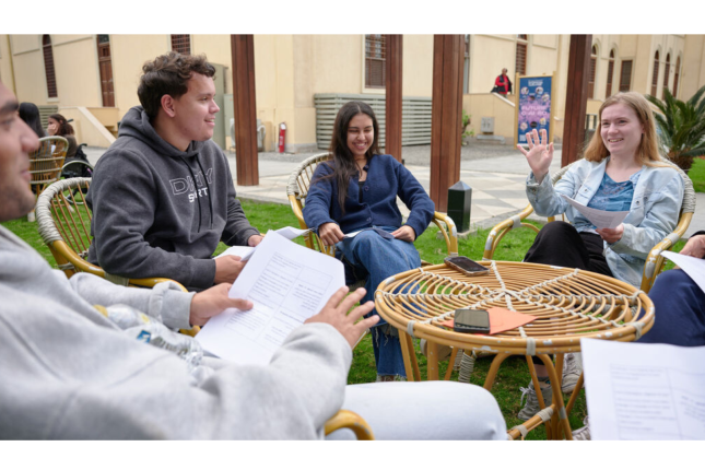 Students hold paper pamphlets while seated in chairs outside and talk with each other