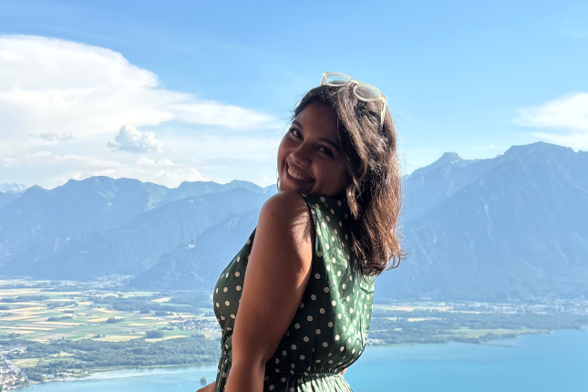 A girl standing in a balcony and behind her is a view of mountains, the sea, and greenery