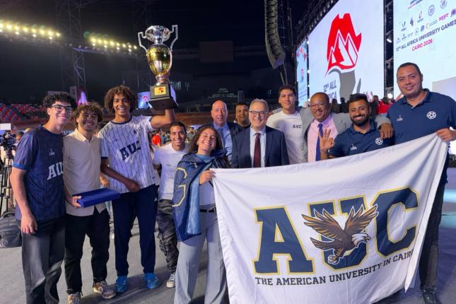 A group of AUC athletes stand with President Ahmad Dallal and Dean of Students Yorgen Marcel smiling, as one of them holds up the cup