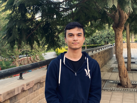 Young man with short dark hair stands in front of trees on a college campus, wearing a navy hoodie