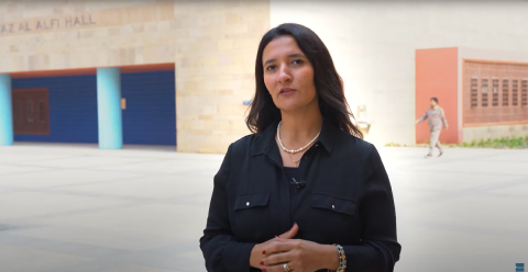 A woman standing in front of a building, she is wearing a dark green blouse