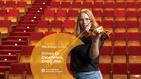 A female playing the violin in front of red stairs, text reads "The Envoy: Judith. Driven by Excellence. Every Day. The American University in Cairo"