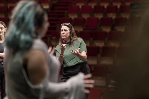 A woman in a green shirt stands in front of red theater chairs, a student in a gray shirt in the foreground