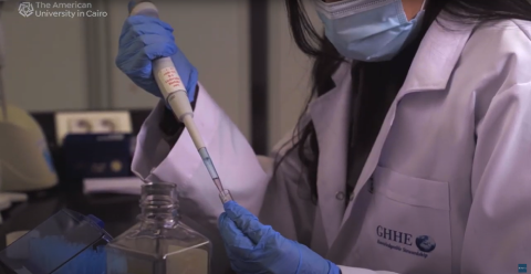 A woman is wearing a lab coat and a mask in a lab and she is holding a test tube. Text: GHHE