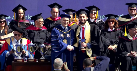A man is shaking hands with a graduate student who is holding a winning cup. There are people sittng behind them and the are all wearing caps and gowns