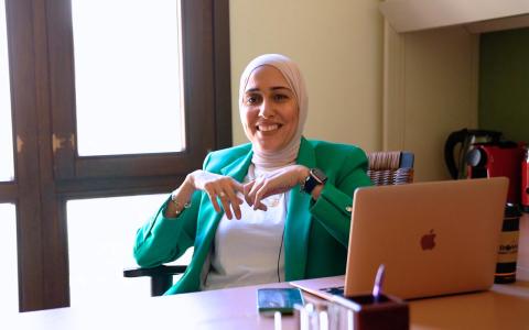 Woman in green jacket and white hijab sits at a desk smiling