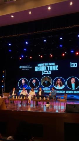 Panel sitting on stage. Text: The American University in Cairo. Sharks take on AUC. Shark Tank مصر