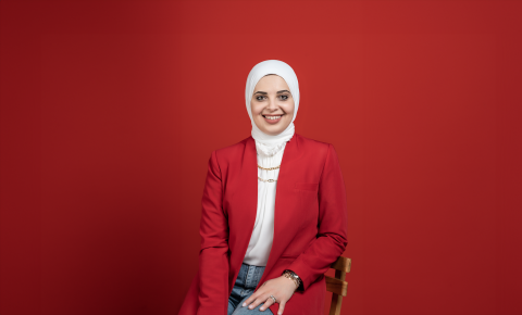 Woman in a red suit jacket and white hijab smiles in front of a red background
