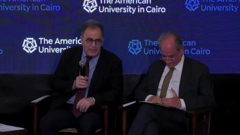 Two men sitting down, one of them is holding a microphone and talking in it