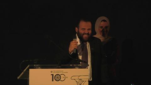 A man is standing at a podium talking into a microphone and a veiled woman is standing behind him
