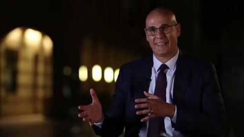 A man is talking in a dimmed setting. He is wearing glasses and a suit